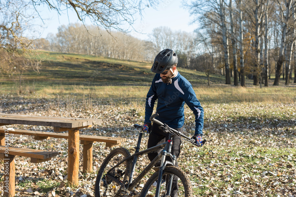 Fototapeta premium Cyclist in pants and fleece jacket on a modern carbon hardtail bike with an air suspension fork rides off-road. The guy is resting on a bench in the autumn park.
