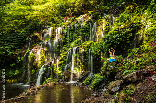 Woman sitting on the rock, practicing yoga. Young woman raising arms with namaste mudra near waterfall. Banyu Wana Amertha waterfall, Bali. View from back. photo