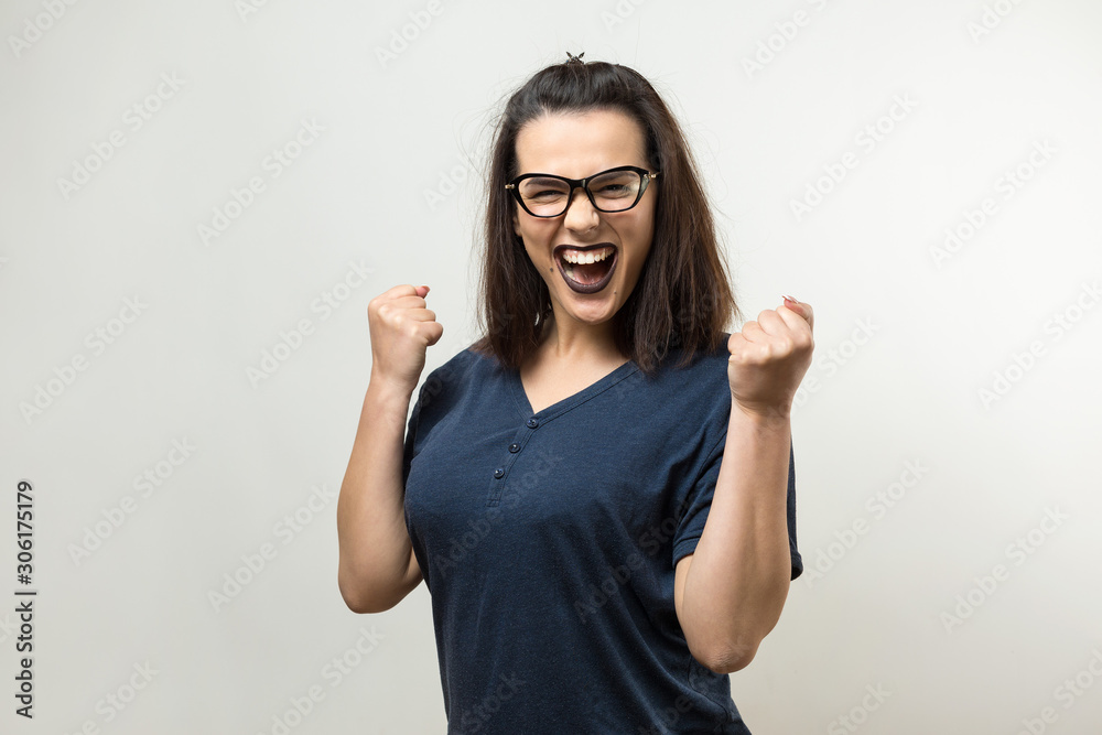 Happy young caucasian femalein in a blue shirt with glasses lifts a clenched fist up for joy. Success and happiness of the work. Successful person, victory! Happy young caucasian femalein in a blue shirt with glasses lifts a clenched fist up for joy. Success and happiness of the work. Successful person, victory!