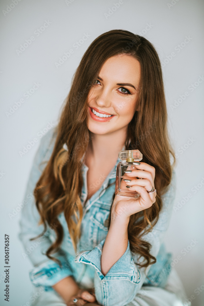 Portrait of young beautiful woman with long hairstyle, wearing blue denim jacket, holding bottle of perfume Portrait of young beautiful woman with long hairstyle, wearing blue denim jacket, holding bottle of perfume