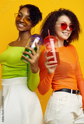 Two young beautiful hipster girls in colorful summer clothes with cocktails