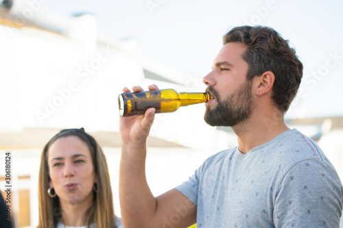 Focused bearded guy enjoying beer with friends on outdoor terrace. Young man and woman in casual meeting outside. Beer party on rooftop concept