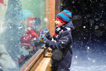 Cute little healthy school kid boy on Christmas market. Funny happy child in fashion winter clothes making window shopping decorated with gifts, xmas tree. Snow falling down, snowfall