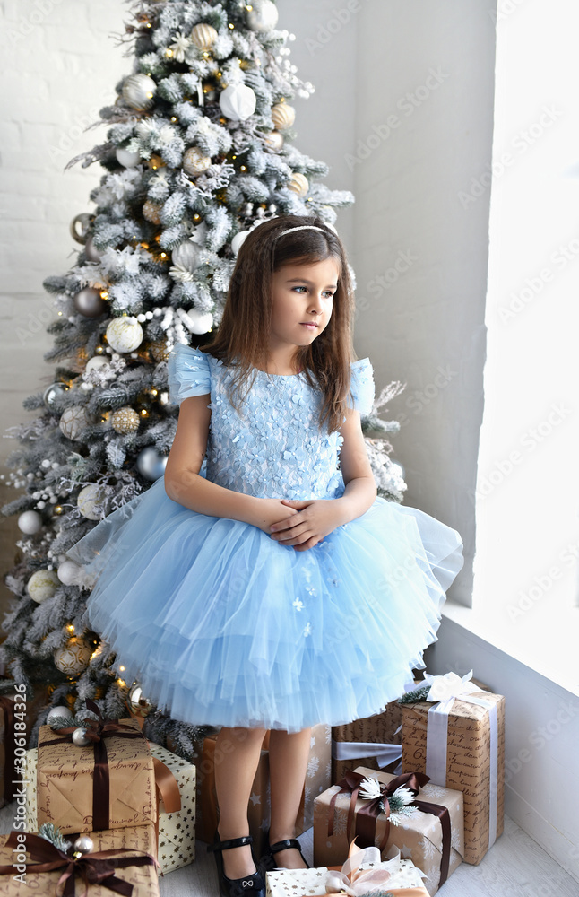 Little girl near fir-tree with Christmas gifts in white and gold holiday interior. Christmas celebration. Child Girl weared in blue dress near a New Year's gifts on the floor. New year eve Little girl near fir-tree with Christmas gifts in white and gold holiday interior. Christmas celebration. Child Girl weared in blue dress near a New Year's gifts on the floor. New year eve