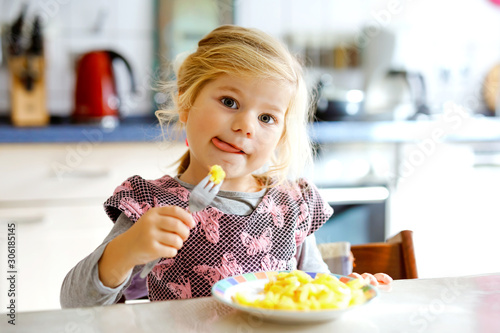 Lovely toddler girl eating healthy fried potatoes for lunch. Cute happy baby child in colorful clothes sitting in kitchen of home  daycare or nursery. Kid eats vegetables.