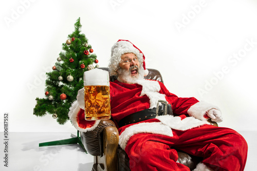 Santa Claus drinking beer near the Christmas tree, congratulating, looks drunk and happy. Caucasian male model in traditional costume. New Year 2020, gifts, holidays, winter mood. Copyspace for your