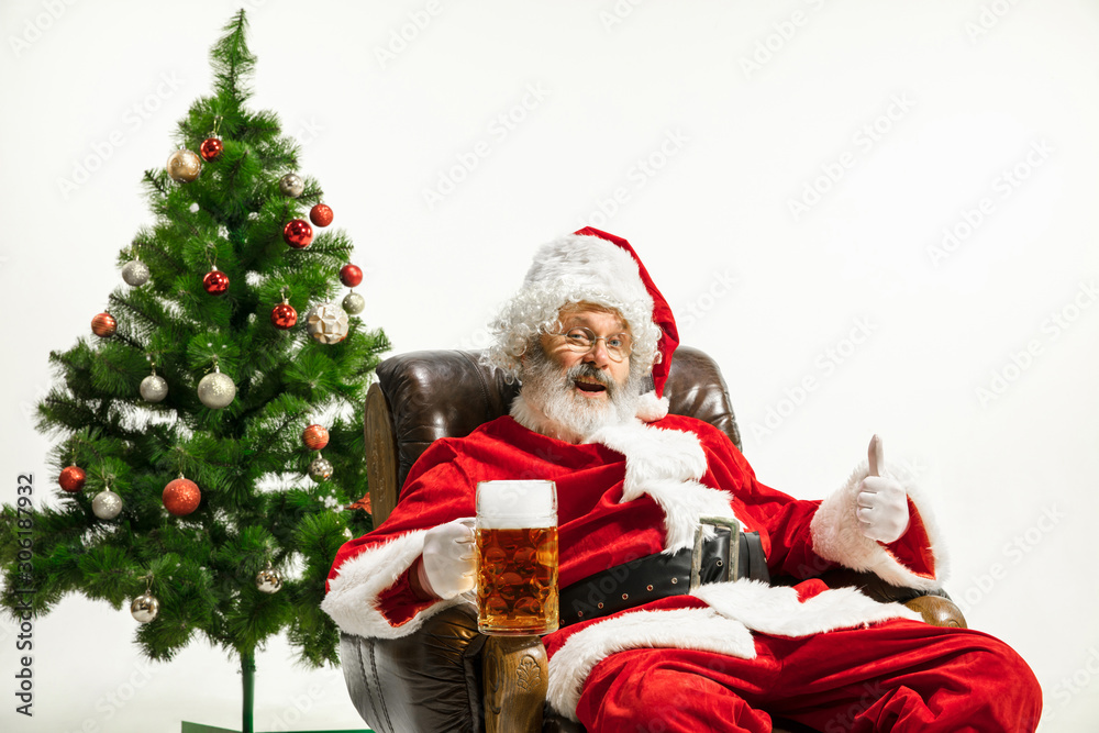 Santa Claus drinking beer near the Christmas tree, congratulating, looks drunk and happy. Caucasian male model in traditional costume. New Year 2020, gifts, holidays, winter mood. Copyspace for your
