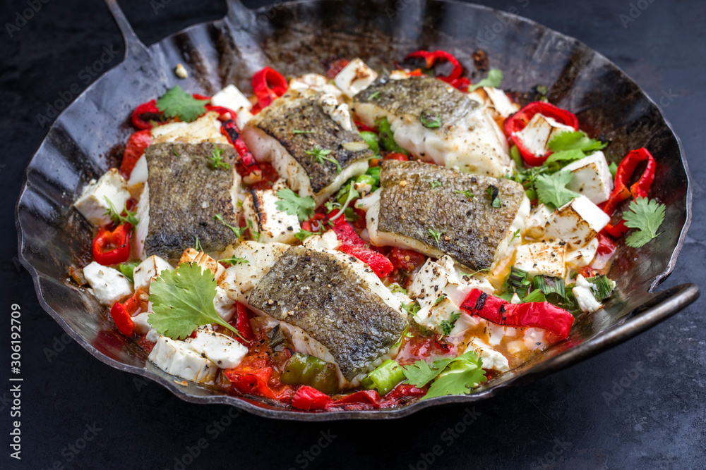 Traditional fried Greek cod fish filet casserole with feta cheese and vegetable as closeup in a rustic wrought iron pan