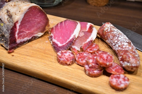 Slices of raw salted homemade ham or prosciutto and sausage on a wooden board with knife