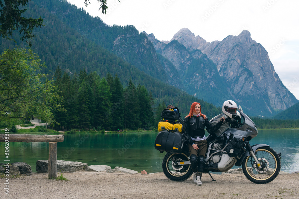 Girl motobiker in protective turtle jacket stands on the beach with tourer motorcycle. Extreme travel vacation, motorcyclist adventure lifestyle. Toblacher See, (Italian: Lago di Dobbiaco) Italy.