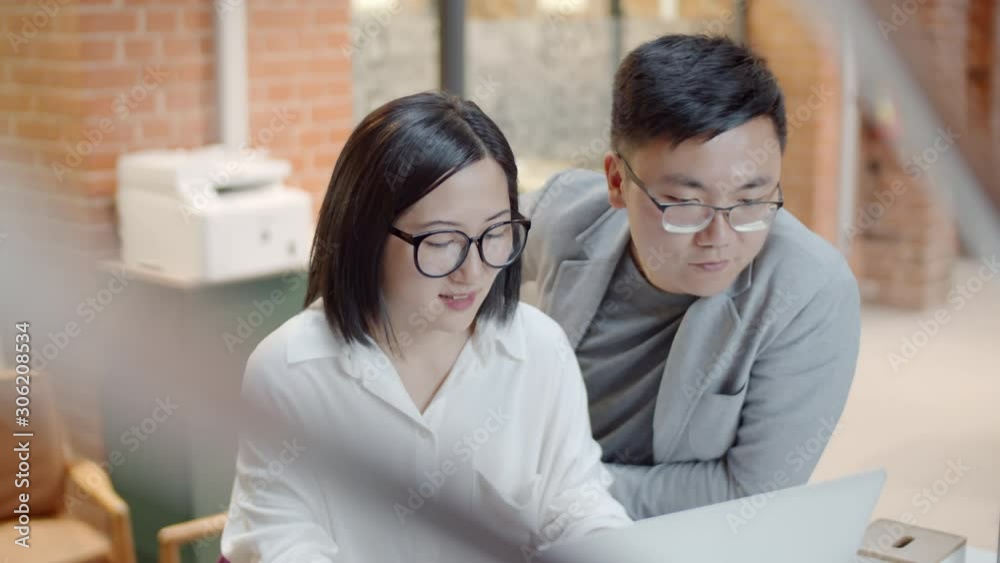 Medium shot of Asian man and woman sitting together at desk, looking at computer screen and having discussion Medium shot of Asian man and woman sitting together at desk, looking at computer screen and having discussion