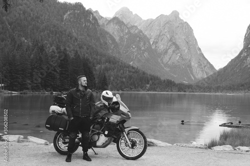Handsome man motorcyclist with touring motorcycle on beach. Alpine mountains and lake on background. Biker lifestyle, world traveler. Toblacher See, (Lago di Dobbiaco) Italy. Black and white