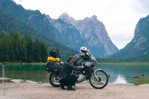 Handsome man motorcyclist repairs, does something on a motorcycle on beach. Alpine mountains and lake on background. Biker lifestyle, world traveler. Toblacher See, (Lago di Dobbiaco) Italy.