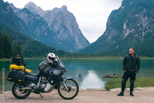 Handsome man motorcyclist with touring motorcycle on beach. Alpine mountains and lake on background. Biker lifestyle, world traveler. Toblacher See, (Lago di Dobbiaco) Italy. copy space in center