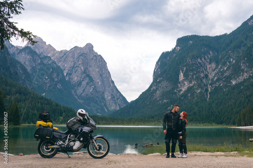 Motorcyclists couple stand on beach with touring notorcycle. Lake and Alpine mountains on background. Destination concept. Happy together. Copy space. Toblacher See, (Lago di Dobbiaco) Italy