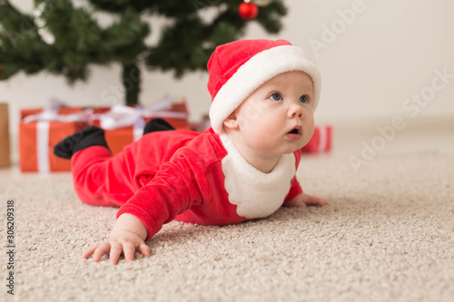 Cute baby girl wearing santa claus suit crawling on floor over Christmas tree. Holiday season.
