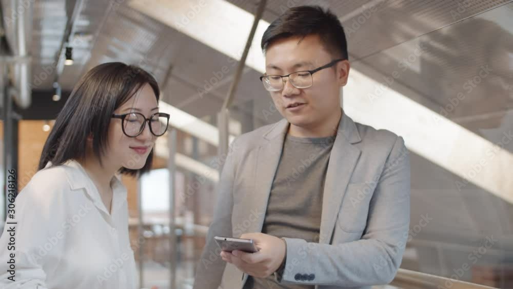 Medium shot of young Asian businessman with telephone in his hands chatting with female colleague standing nearby Medium shot of young Asian businessman with telephone in his hands chatting with female colleague standing nearby