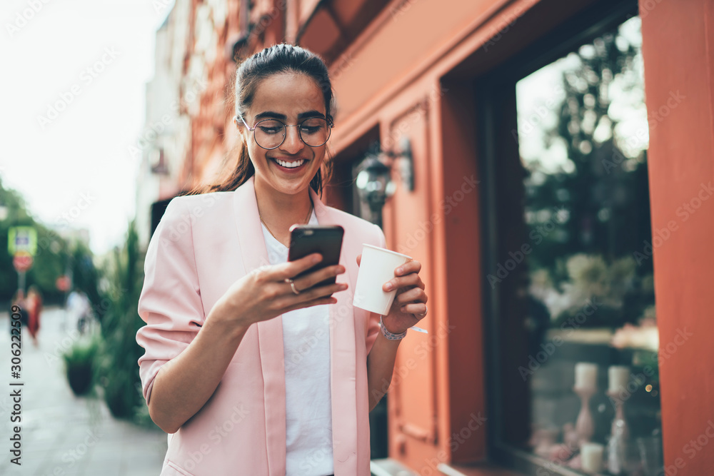 Cheerful woman in big classic glasses for provide eyes protection typing text message during time for exploring new city on vacations using public internet connection on modern cellphone gadget