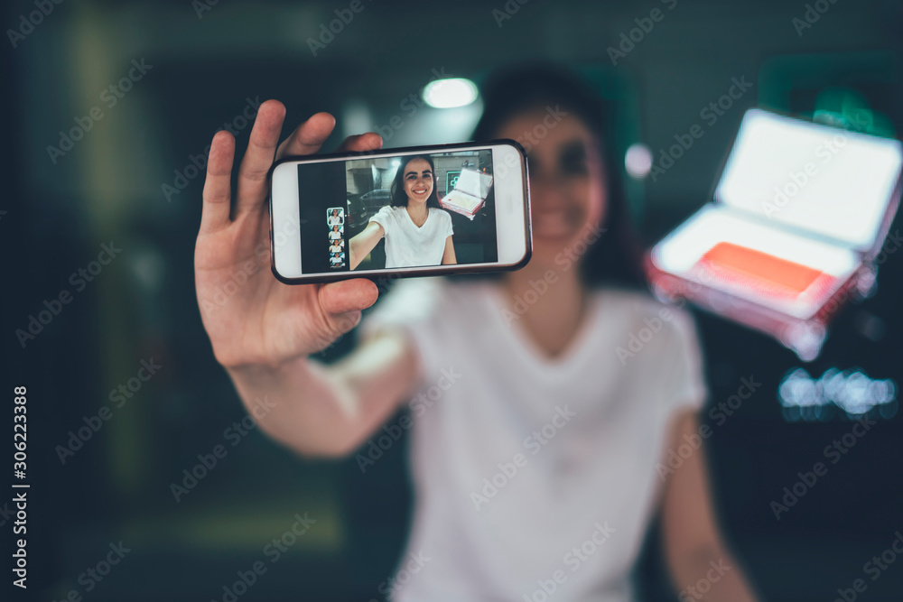 Selective focus on modern cellphone gadget holding positive millennial hipster girl, woman using main camera on smartphone device for photographing herself and attracting followers with publication