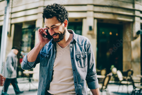 Cheerful bearded hipster blogger laughing during phone conversation with mobile operator on street.Positive young man dressed in denim shirt strolling near mall and calling on cellular