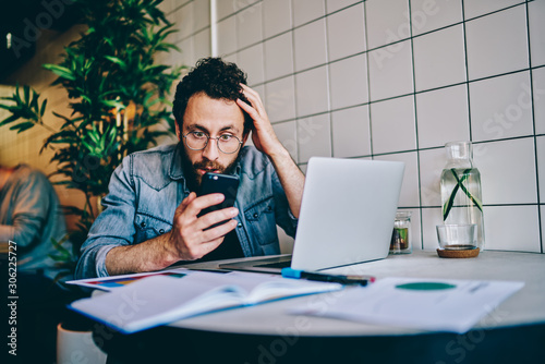 Excited bearded hipster blogger holding head with hand while reading shocked notification on smartphone during distance job at laptop.Amazed young man watching shocked video on web page on cellular