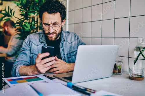 Caucasian hipster guy confused with getting message with bed news on smartphone during remote job in coffee shop, serious young male blogger disappointed with banking app failure on mobile phone .