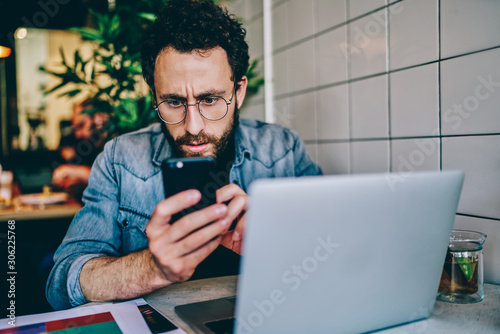 Concentrated male blogger holding mobile phone close to face read carefully message, serious caucasian hipster guy confused with news from social networks checking mail on smartphone on remote job.