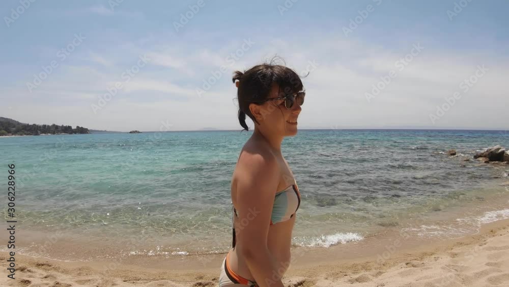 A young brunette girl in swimwear smiling and walking by the sea on a sandy beach in Greece on a sunny summer day. A young brunette girl in swimwear smiling and walking by the sea on a sandy beach in Greece on a sunny summer day.