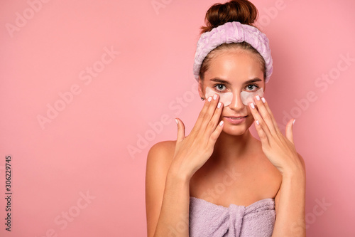 A girl with clean skin stands on a pink background in a towel and with her fingers straightens patches under her eyes.