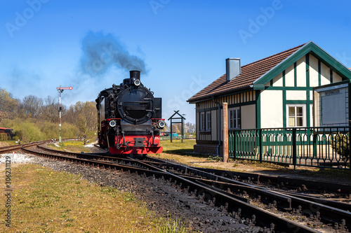Dampflok, Schmalspurbahn bei einfahrt in Bahnhof mit Stellwerkhäuschen und Weichen vor blauem Himmel mit dampf