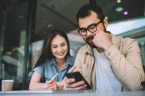 Happy woman watching funny video on asian boyfriend's mobile phone spending free time together in coffee shop, hipster guys checking nes update for application on smartphone via 4G internet. photo