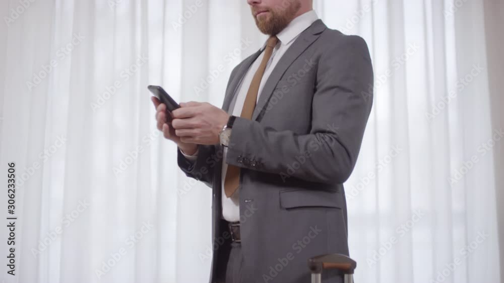 Tilt down shot of successful businessman in suit standing before window in hotel room and typing on mobile phone, then taking his suitcase and leaving Tilt down shot of successful businessman in suit standing before window in hotel room and typing on mobile phone, then taking his suitcase and leaving