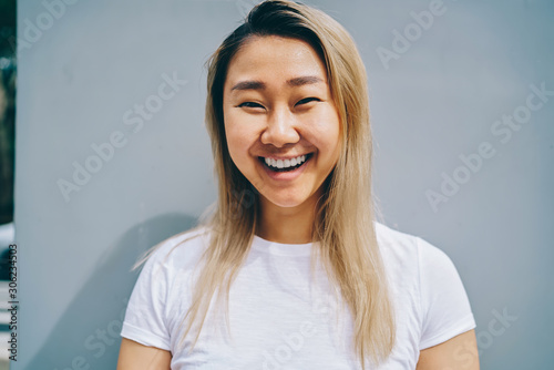 Half length portrait of cute asian girl having fun during free time laughing posing on blue background, cheerful blonde asian woman in shirt with copy space for brand name or label looking at camera