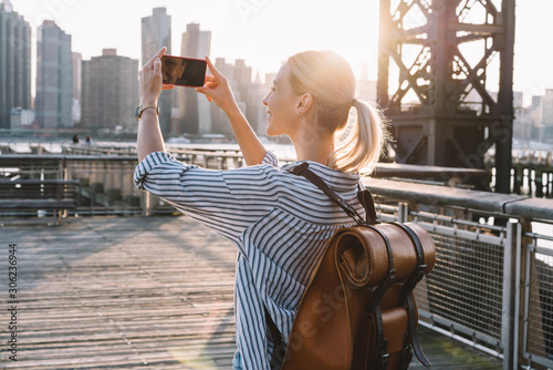 Back view of millennial woman shooting video of New York landscape using smartphone for clicking pictures, positive female tourist testing cellphone camera for taking photos of city in sunrise photo