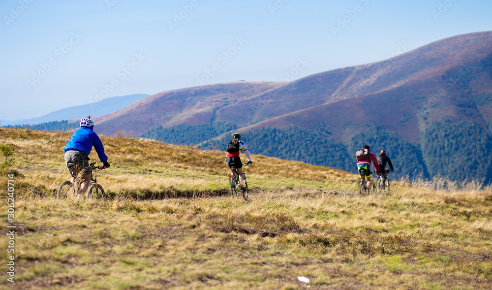 Obraz premium Group of unrecognizable tourists friends ride bicycles along top of mountains road in scenic green valley. Mountain bikers cruise through picturesque nature on sunny day.