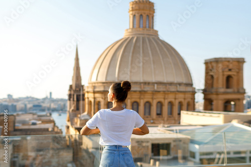 A brunette woman stands on a balcony at a height against the background of the city and the church with a dome. Focus on the woman. Back view photo