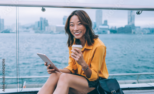 Portrait of cheerful hipster girl with modern digital tablet and coffee to go rejoicing outdoors enjoying Chinese vacations and time for exploring Hong Kong metropolis using 4g wireless for research #306242936