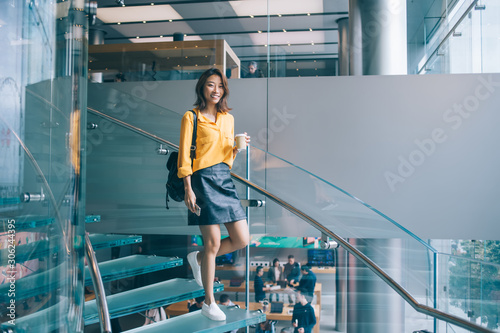 Pretty Asian woman going down stairs with hot drink and cellphone