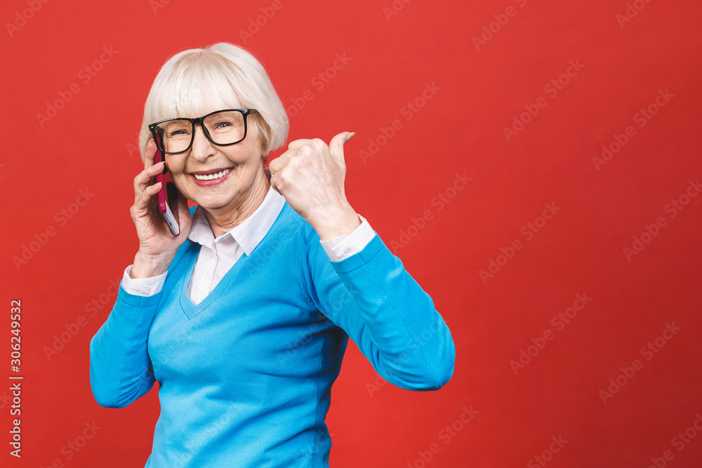 Senior woman using smartphone standing over isolated red background pointing and showing with thumb up to the side with happy face smiling Senior woman using smartphone standing over isolated red background pointing and showing with thumb up to the side with happy face smiling