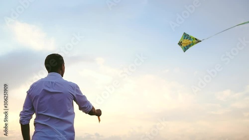 young man in white shirt flies colorful kite in sky with light clouds at sunset backside low angle shot slow motion