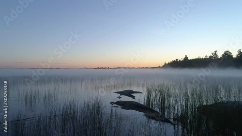 4K aerial flight over rocks and reeds with fog in the distance at a lake on a misty fall morning in Northwest Ontario, Canada.