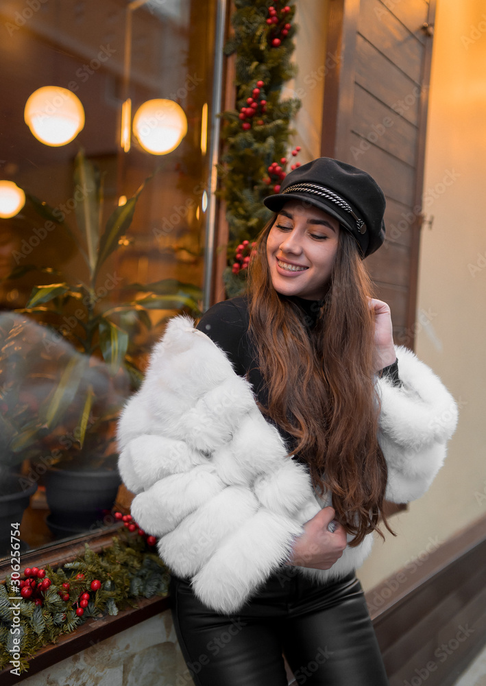 Beautiful young caucasian woman wearing natural short white fur coat posing for a portrait near Christmas showcase. Christmas decor. Fashion. Style. Winter clothes made of natural fur. Vertical