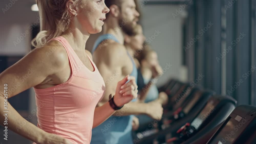 Fit Athletic Woman Running on the Treadmill, Doing Her Fitness Exercise. Muscular Women and Men Actively Training in the Modern Gym. Sports People Workout in Fitness Club. Side View Slow Motion
