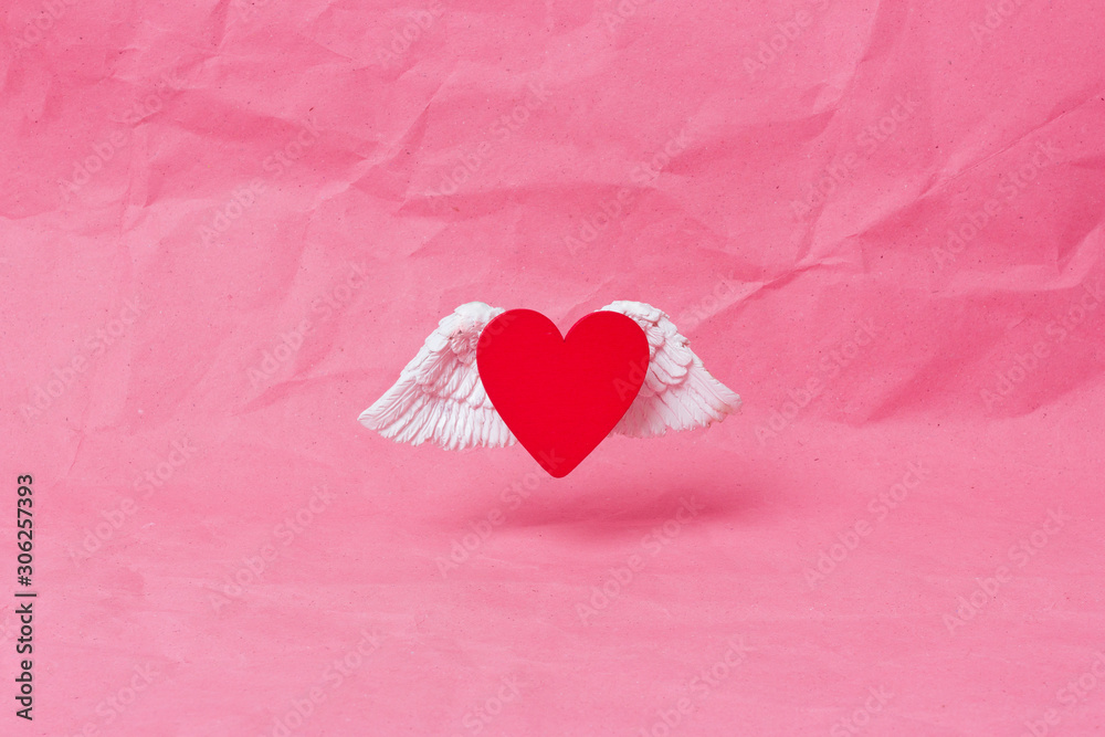 Fototapeta premium Happy valentines day banner. A red wooden heart with white voluminous wings soars over a pink crumpled paper background. Minimalism. Place for text