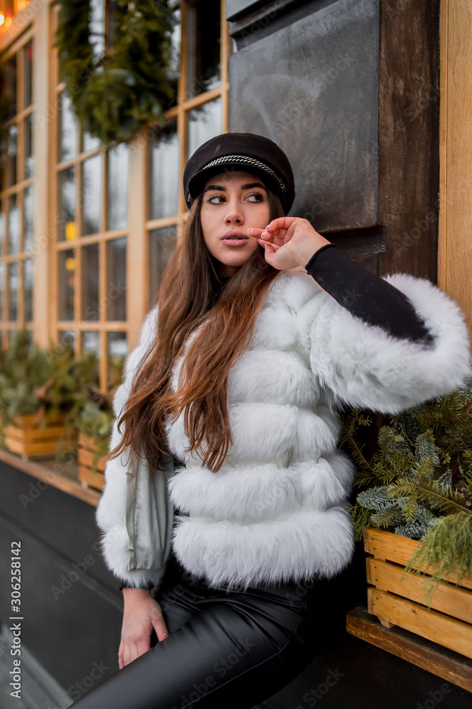 Beautiful young caucasian woman wearing natural short white fur coat posing for a portrait near Christmas showcase. Christmas decor. Fashion. Style. Winter clothes made of natural fur. Vertical