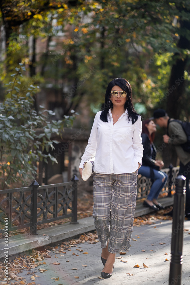 Autumn fashion outdoor. The girl in fashionable stylish white shirt , pants and sunglasses, autumnal lifestyle on the background of blurry yellow-green trees in the park. Vertical Autumn fashion outdoor. The girl in fashionable stylish white shirt , pants and sunglasses, autumnal lifestyle on the background of blurry yellow-green trees in the park. Vertical