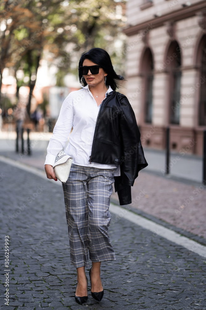 Fototapeta premium Autumn fashion outdoor. The girl in fashionable stylish white shirt , pants, jacket and sunglasses, autumnal lifestyle on the background of blurry yellow-green trees in the park. Vertical