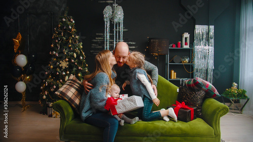 Christmas concept - family sitting on the sofa - dad giving others a gift - his wife and daughter kissing him in cheeks © KONSTANTIN SHISHKIN