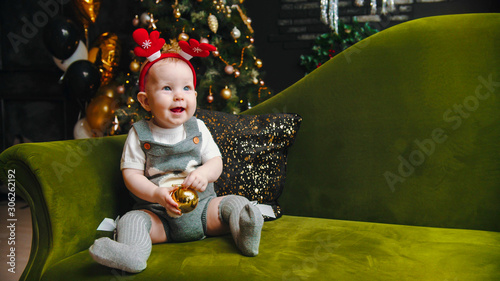 Christmas concept - smiling baby is sitting on sofa and playing with christmas ball