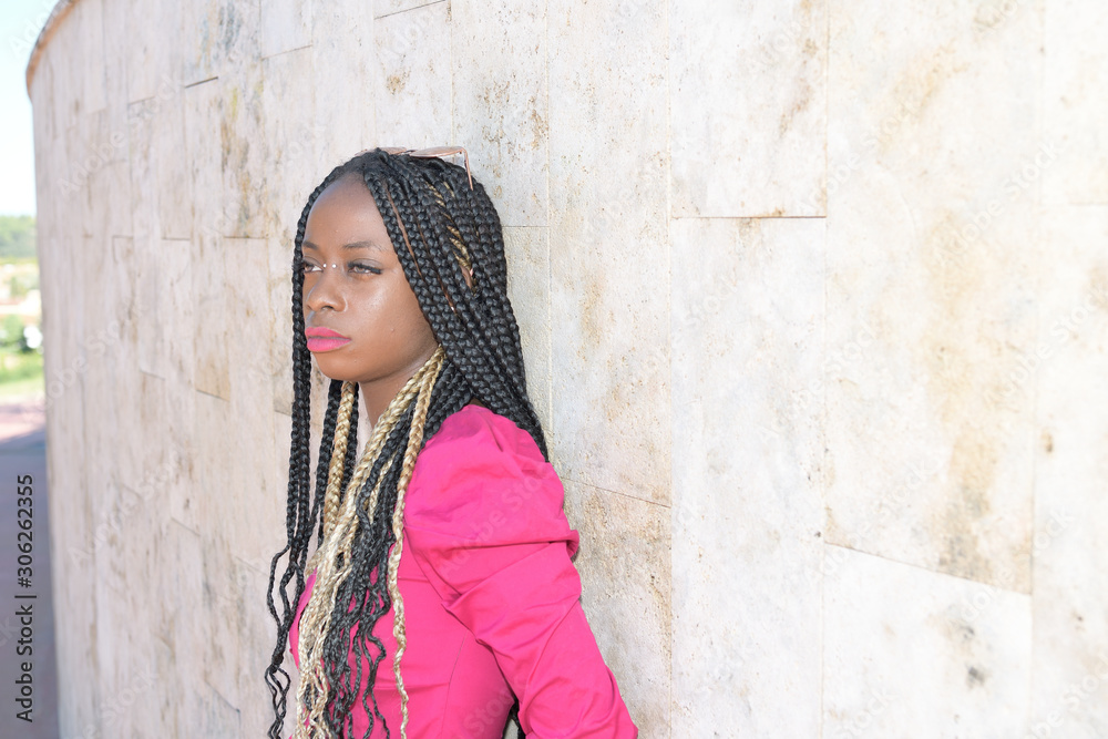 Fototapeta premium Afro-style black girl with long black and blondes braids wearing sunglasses rests on a wall and daydreams